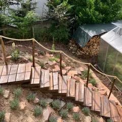 Eine einfache Gartentreppe aus Holz mit Seilgeländer, die sich den Hang hinunter schlängelt. Daneben ein Gewächshaus und ein Holzstapel.
