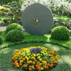 Ein Grab wird liebevoll bepflanzt mit bunten Blumen und immergrünen Buchsbäumen. Ein runder Stein mit Gravur steht im Hintergrund.