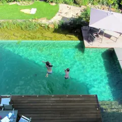 Kinder springen in einen kristallklaren Pool, der Teil eines Gartens mit einem **schwimmteich anlegen** ist. Eine Holzterrasse mit Sonnenschirm lädt zum Entspannen ein.