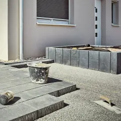 Gartenbauarbeiten: Pflastersteine liegen bereit, Werkzeuge wie Gummihammer und Kelle sind platziert. Ein Hochbeet aus Betonpalisaden wird gerade gesetzt.