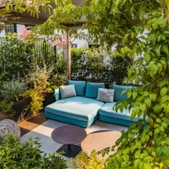 Traumhafte dachterrasse ideen: Eine türkise Lounge-Couch unter üppigem Grün, mit zarten rosa Blüten und zwei runden Tischen.
