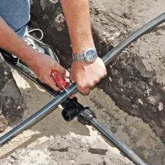 Mann verlegt eine neue wasserleitung im garten. Mit einem Rohrschneider wird das schwarze Rohr zugeschnitten, um es an ein T-Stück anzuschließen.