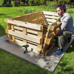 Mann baut ein schmales Hochbeet aus Paletten im Garten. Werkzeug und Schrauben liegen bereit.
