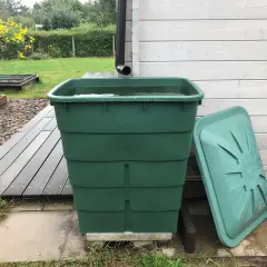 Grüner Regentank fängt Wasser auf. Daneben liegt der Deckel. Im Hintergrund ein Garten mit gelben Blumen.