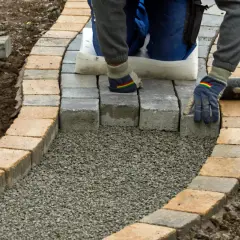 Arbeiter legt mit Handschuhen und Werkzeug einen neuen Gartenweg an, Stein für Stein.
