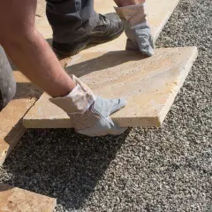 Arbeiter verlegt mit Handschuhen eine helle Steinplatte auf Kies. Präzises Arbeiten beim Platten verlegen für eine schöne Terrasse.