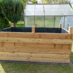 Der **Aufbau** eines **Hochbeets** aus Holzbrettern im Garten. Im Hintergrund ein Gewächshaus und grüne Büsche.