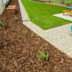 Rindenmulch gegen Unkraut schützt junge Pflanzen am Zaun. Daneben ein gepflegter Rasen und eine Terrasse mit Gartenmöbeln.
