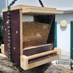 Selbstgebautes vogelfutterhaus aus Holz und Kunststoff, gefüllt mit Körnern, hängt an einer Kette an einem alten Holzpfosten.