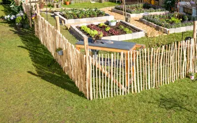 Ein rustikaler Staketenzaun wird befestigt, um einen gepflegten Gemüsegarten mit Hochbeeten und einer Sitzgelegenheit abzugrenzen.