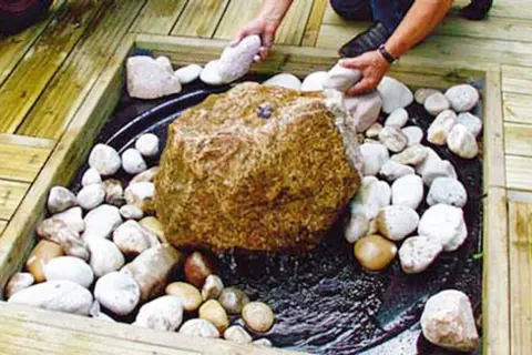 Beim **gartenbrunnen selber bauen** wird ein großer Stein mit kleineren Steinen umgeben. Wasser plätschert.