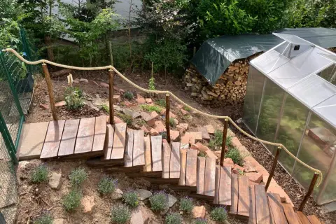 Eine einfache Gartentreppe aus Holz mit Seilgeländer, die sich den Hang hinunter schlängelt. Daneben ein Gewächshaus und ein Holzstapel.
