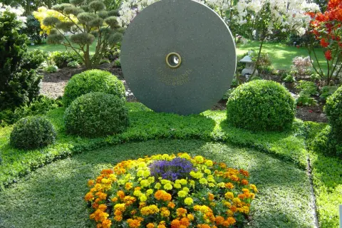 Ein Grab wird liebevoll bepflanzt mit bunten Blumen und immergrünen Buchsbäumen. Ein runder Stein mit Gravur steht im Hintergrund.