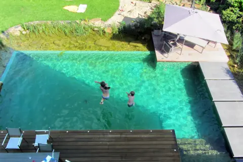Kinder springen in einen kristallklaren Pool, der Teil eines Gartens mit einem **schwimmteich anlegen** ist. Eine Holzterrasse mit Sonnenschirm lädt zum Entspannen ein.