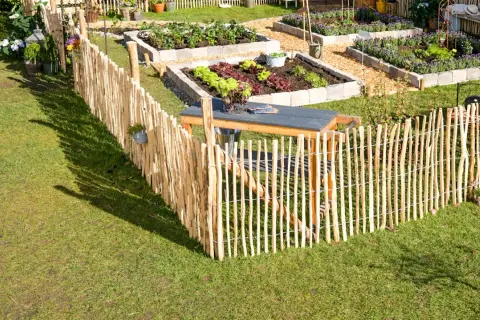 Ein rustikaler Staketenzaun wird befestigt, um einen gepflegten Gemüsegarten mit Hochbeeten und einer Sitzgelegenheit abzugrenzen.