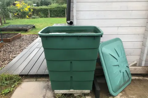 Grüner Regentank fängt Wasser auf. Daneben liegt der Deckel. Im Hintergrund ein Garten mit gelben Blumen.
