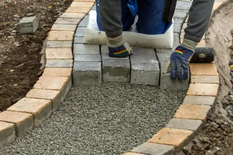 Arbeiter legt mit Handschuhen und Werkzeug einen neuen Gartenweg an, Stein für Stein.