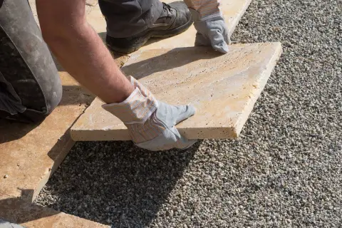 Arbeiter verlegt mit Handschuhen eine helle Steinplatte auf Kies. Präzises Arbeiten beim Platten verlegen für eine schöne Terrasse.