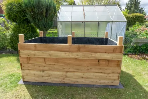 Der **Aufbau** eines **Hochbeets** aus Holzbrettern im Garten. Im Hintergrund ein Gewächshaus und grüne Büsche.