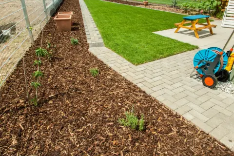 Rindenmulch gegen Unkraut schützt junge Pflanzen am Zaun. Daneben ein gepflegter Rasen und eine Terrasse mit Gartenmöbeln.