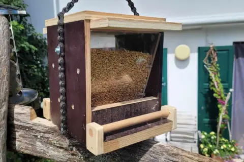 Selbstgebautes vogelfutterhaus aus Holz und Kunststoff, gefüllt mit Körnern, hängt an einer Kette an einem alten Holzpfosten.