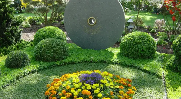 Ein Grab wird liebevoll bepflanzt mit bunten Blumen und immergrünen Buchsbäumen. Ein runder Stein mit Gravur steht im Hintergrund.