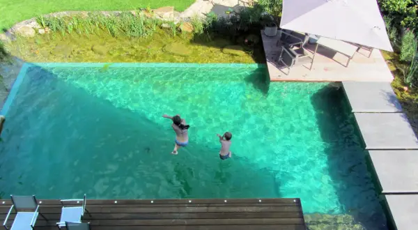 Kinder springen in einen kristallklaren Pool, der Teil eines Gartens mit einem **schwimmteich anlegen** ist. Eine Holzterrasse mit Sonnenschirm lädt zum Entspannen ein.