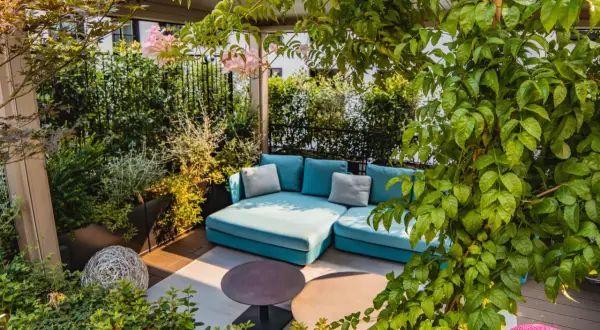 Traumhafte dachterrasse ideen: Eine türkise Lounge-Couch unter üppigem Grün, mit zarten rosa Blüten und zwei runden Tischen.