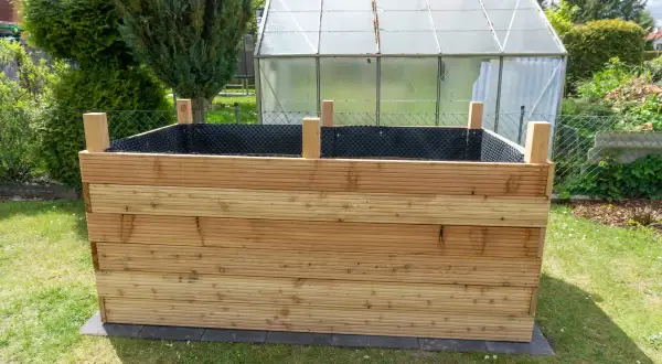 Ein neues Hochbeet bauen: Holzbretter werden zu einem stabilen Rahmen verschraubt, bereit für Erde und Pflanzen. Im Hintergrund ein Gewächshaus.