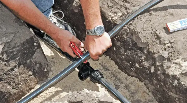 Mann verlegt eine neue wasserleitung im garten. Mit einem Rohrschneider wird das schwarze Rohr zugeschnitten, um es an ein T-Stück anzuschließen.