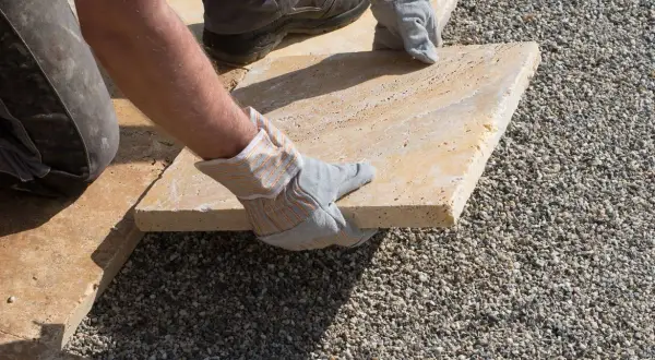 Arbeiter verlegt mit Handschuhen eine helle Steinplatte auf Kies. Präzises Arbeiten beim Platten verlegen für eine schöne Terrasse.
