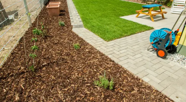Rindenmulch gegen Unkraut schützt junge Pflanzen am Zaun. Daneben ein gepflegter Rasen und eine Terrasse mit Gartenmöbeln.