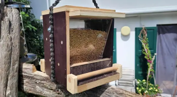 Selbstgebautes vogelfutterhaus aus Holz und Kunststoff, gefüllt mit Körnern, hängt an einer Kette an einem alten Holzpfosten.