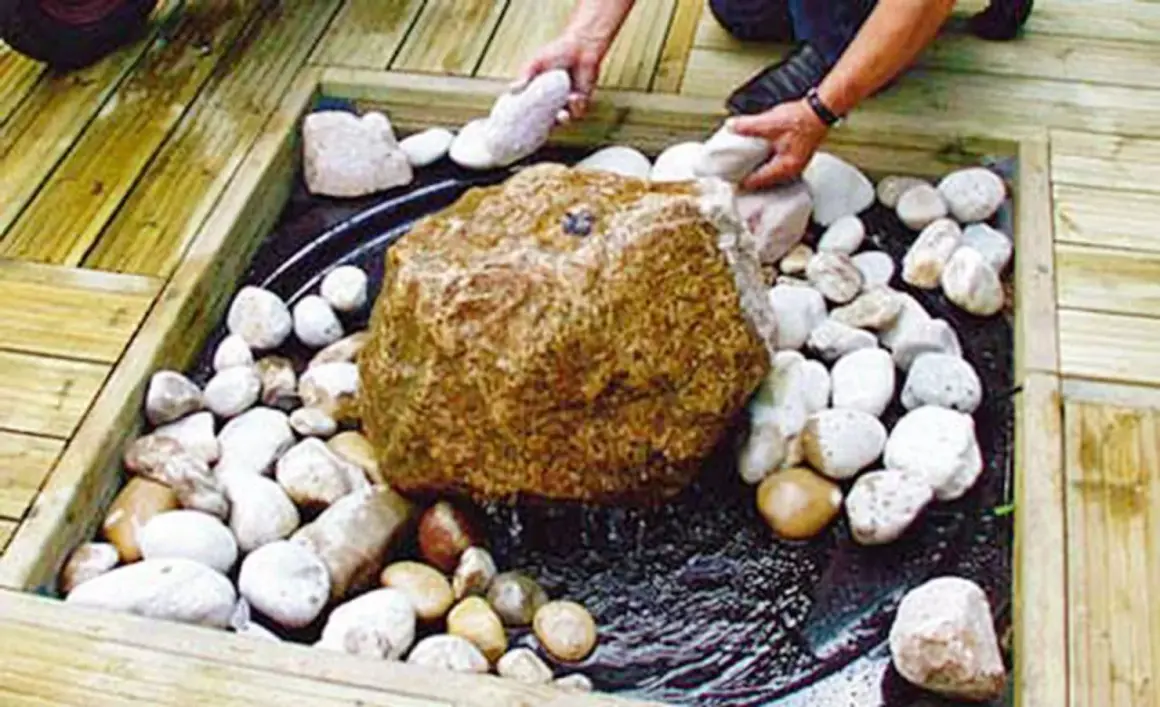 Beim **gartenbrunnen selber bauen** wird ein großer Stein mit kleineren Steinen umgeben. Wasser plätschert.