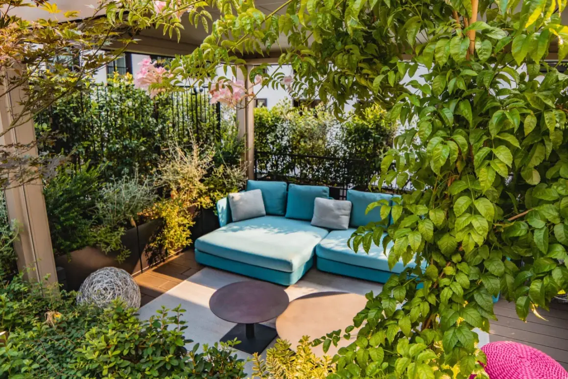 Traumhafte dachterrasse ideen: Eine türkise Lounge-Couch unter üppigem Grün, mit zarten rosa Blüten und zwei runden Tischen.