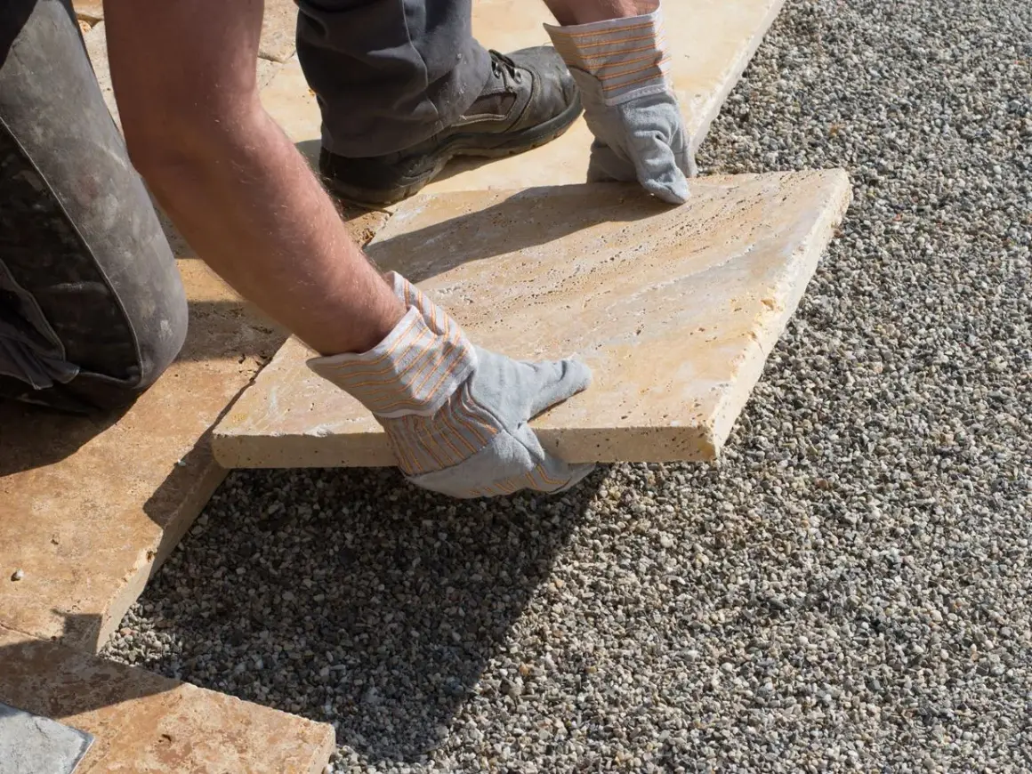 Arbeiter verlegt mit Handschuhen eine helle Steinplatte auf Kies. Präzises Arbeiten beim Platten verlegen für eine schöne Terrasse.