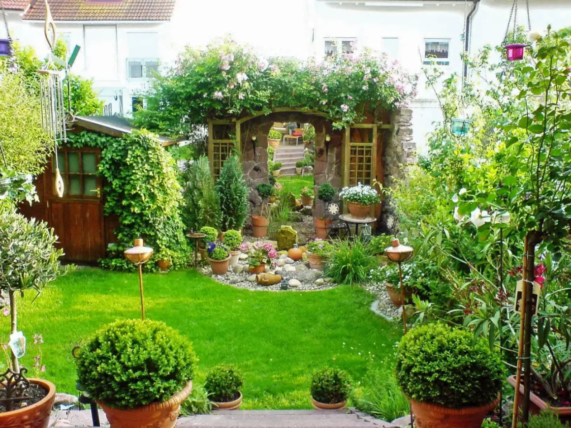 Ein moderner kleiner Garten im Reihenhaus mit Steinbogen, Rosen, Schuppen und viel Grün.