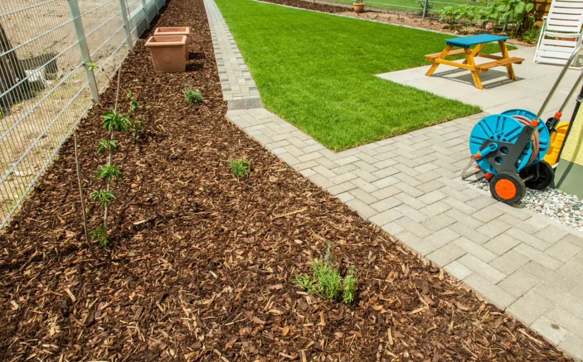 Rindenmulch gegen Unkraut schützt junge Pflanzen am Zaun. Daneben ein gepflegter Rasen und eine Terrasse mit Gartenmöbeln.