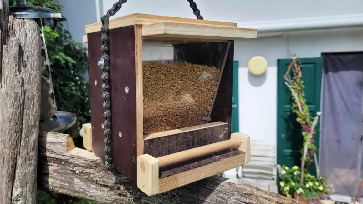 Selbstgebautes vogelfutterhaus aus Holz und Kunststoff, gefüllt mit Körnern, hängt an einer Kette an einem alten Holzpfosten.