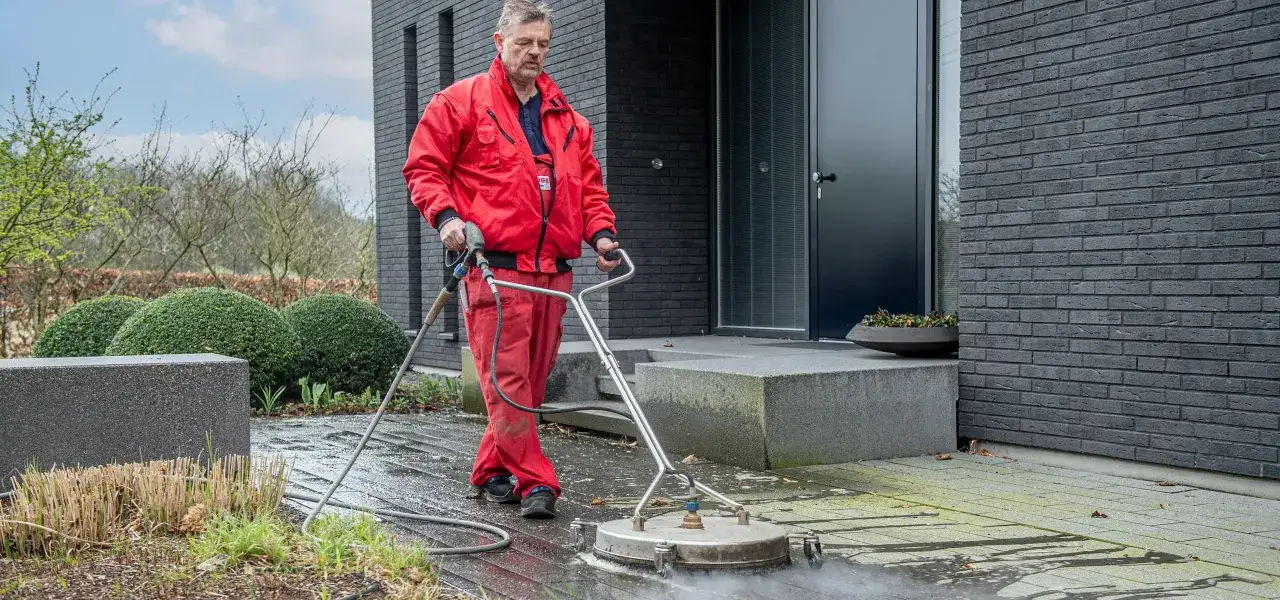 Mann in roter Arbeitskleidung reinigt mit einem Hochdruckreiniger das betonpflaster vor einem modernen Haus.