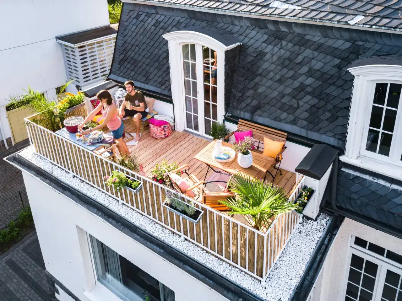 Ein Paar genie&szlig;t ein Grillfest auf einem gem&uuml;tlichen Balkon mit vielen Pflanzen. Coole Gartenideen einfach umgesetzt f&uuml;r entspannte Stunden.