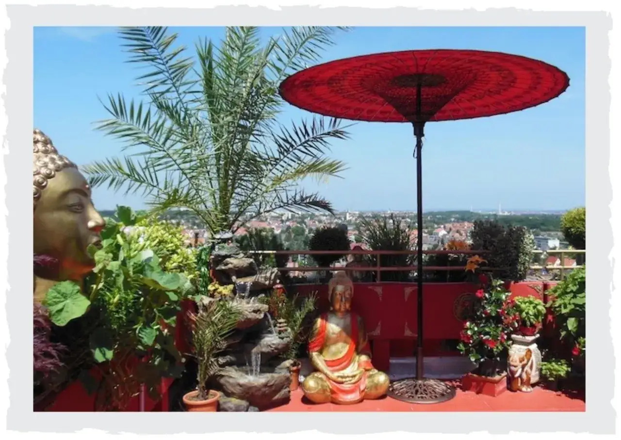 Ein friedlicher, privater japanischer Garten mit Buddha-Statuen, einem kleinen Wasserfall und einem roten Sonnenschirm, der einen Blick auf die Stadt bietet.
