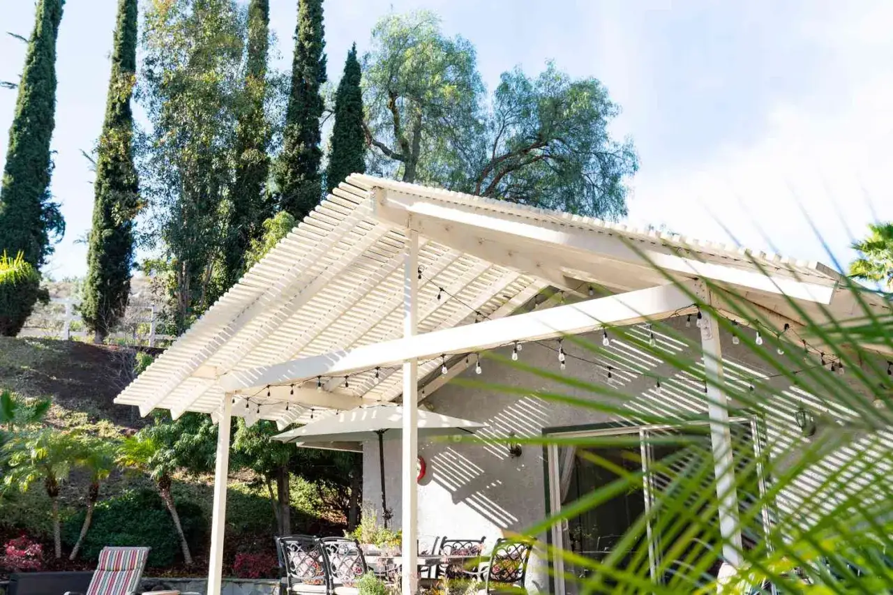 Eine wei&szlig;e Pergola mit Lichterkette schafft sch&ouml;ne Schatten im Garten. Sitzm&ouml;bel und Pflanzen laden zum Verweilen ein.