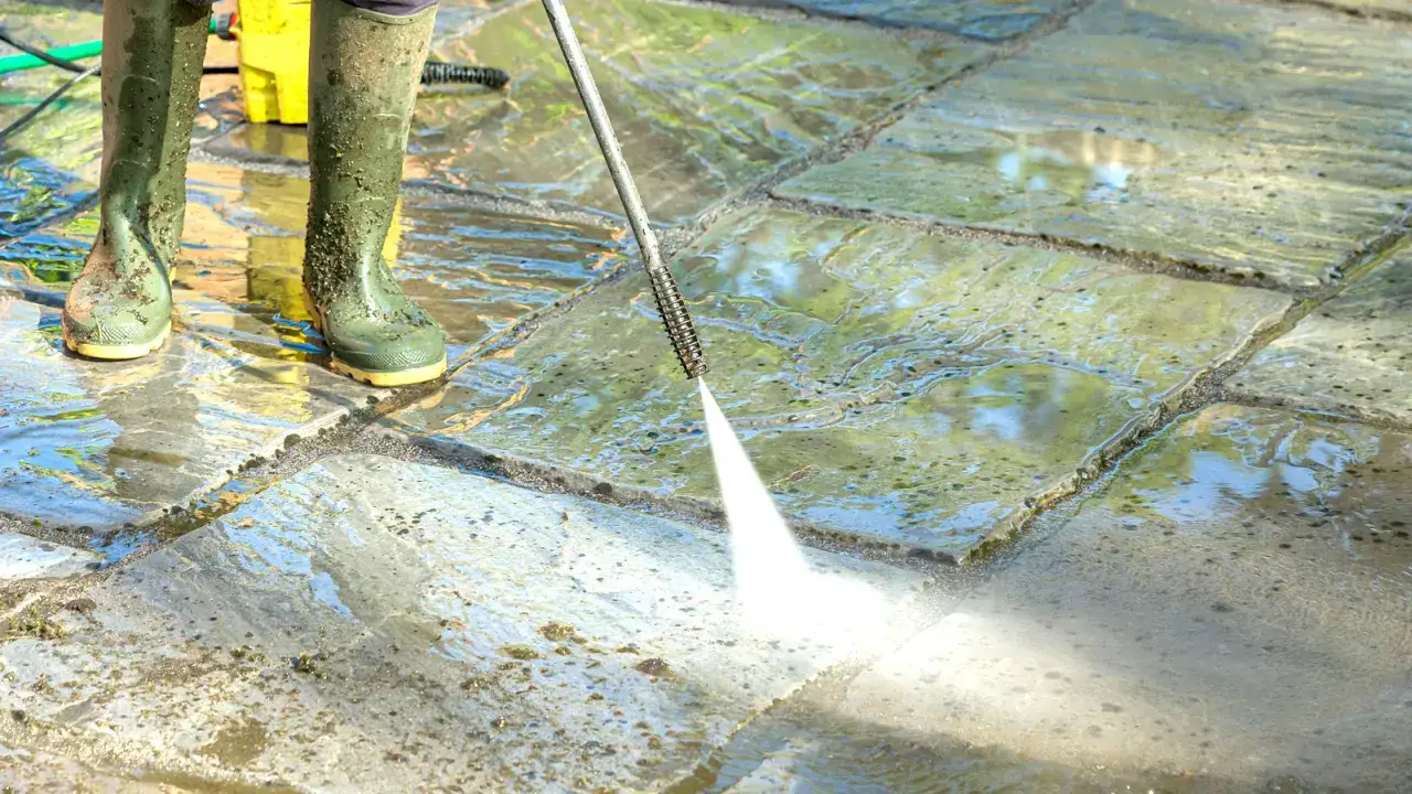 Person in Gummistiefeln reinigt mit Hochdruckreiniger Terrassenplatten. Der Wasserstrahl l&ouml;st Schmutz und Moos.