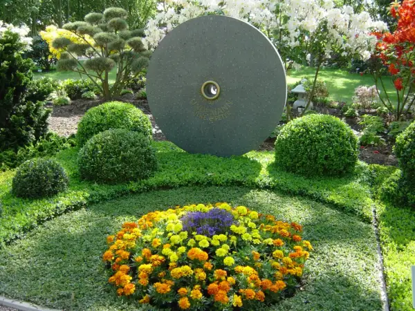 Ein Grab wird liebevoll bepflanzt mit bunten Blumen und immergrünen Buchsbäumen. Ein runder Stein mit Gravur steht im Hintergrund.