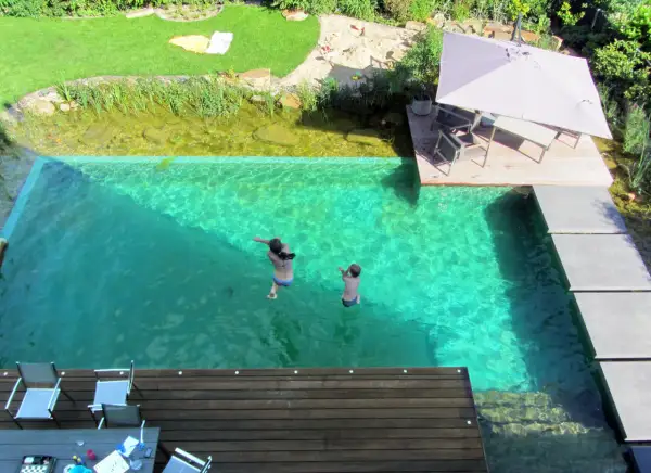 Kinder springen in einen kristallklaren Pool, der Teil eines Gartens mit einem **schwimmteich anlegen** ist. Eine Holzterrasse mit Sonnenschirm lädt zum Entspannen ein.