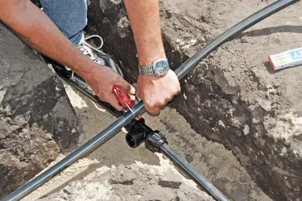 Mann verlegt eine neue wasserleitung im garten. Mit einem Rohrschneider wird das schwarze Rohr zugeschnitten, um es an ein T-Stück anzuschließen.
