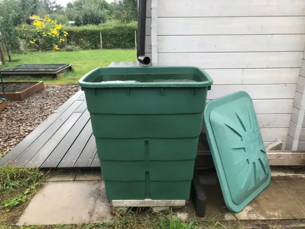 Grüner Regentank fängt Wasser auf. Daneben liegt der Deckel. Im Hintergrund ein Garten mit gelben Blumen.